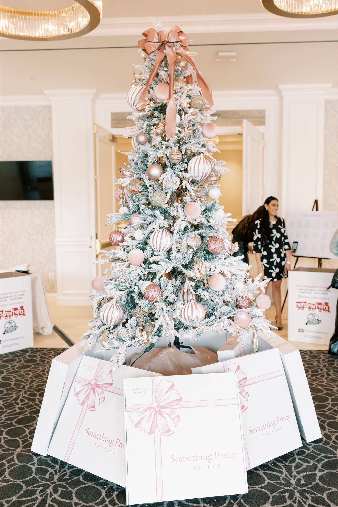Christmas tree with pink ornaments and bow, surrounded by gifts. Indoors, with people in the background.