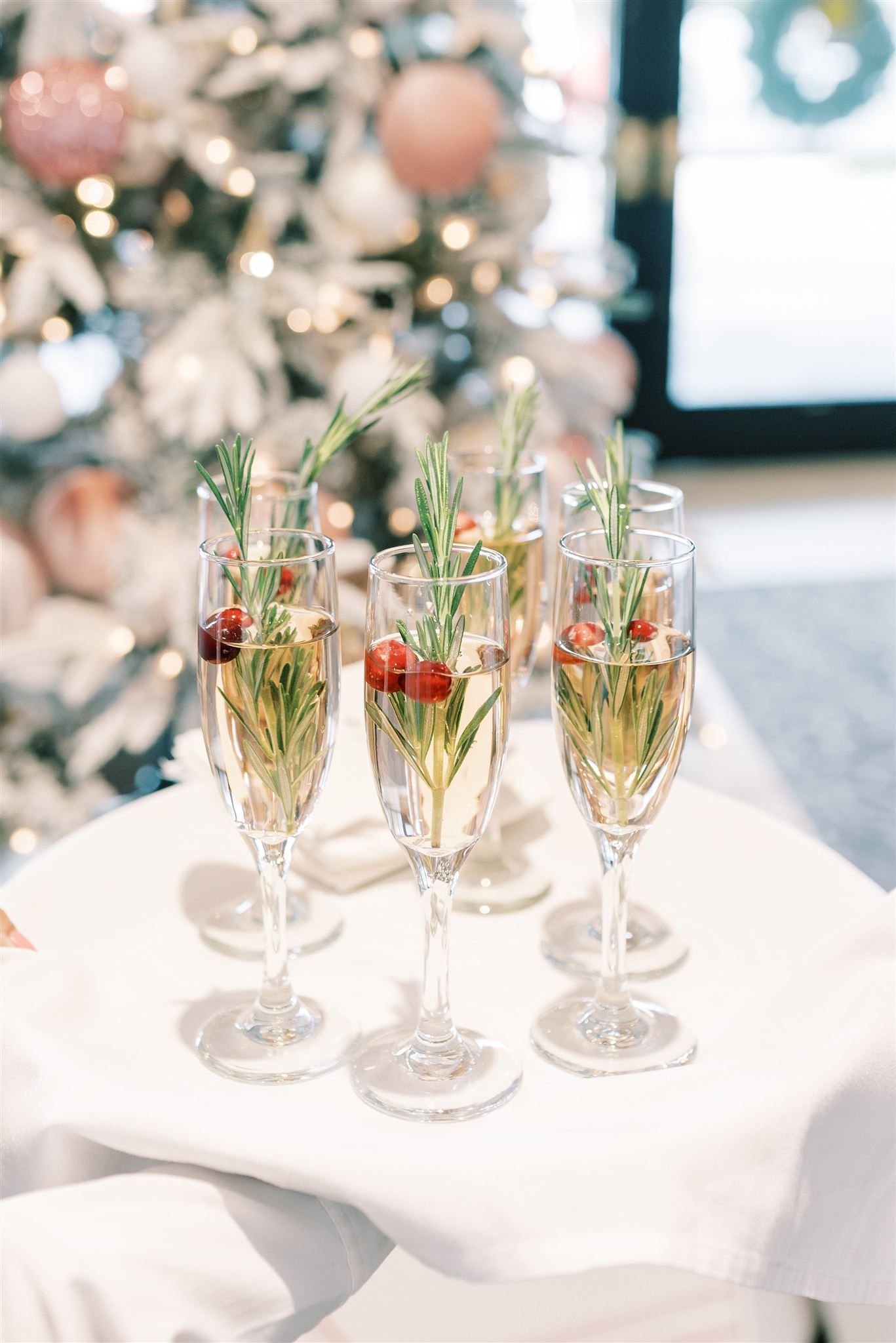 A tray of champagne flutes, each garnished with cranberries and rosemary, near a decorated Christmas tree.