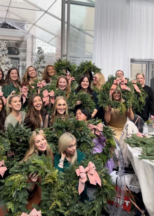 Group of women holding wreaths with pink bows in a workshop setting, smiling.