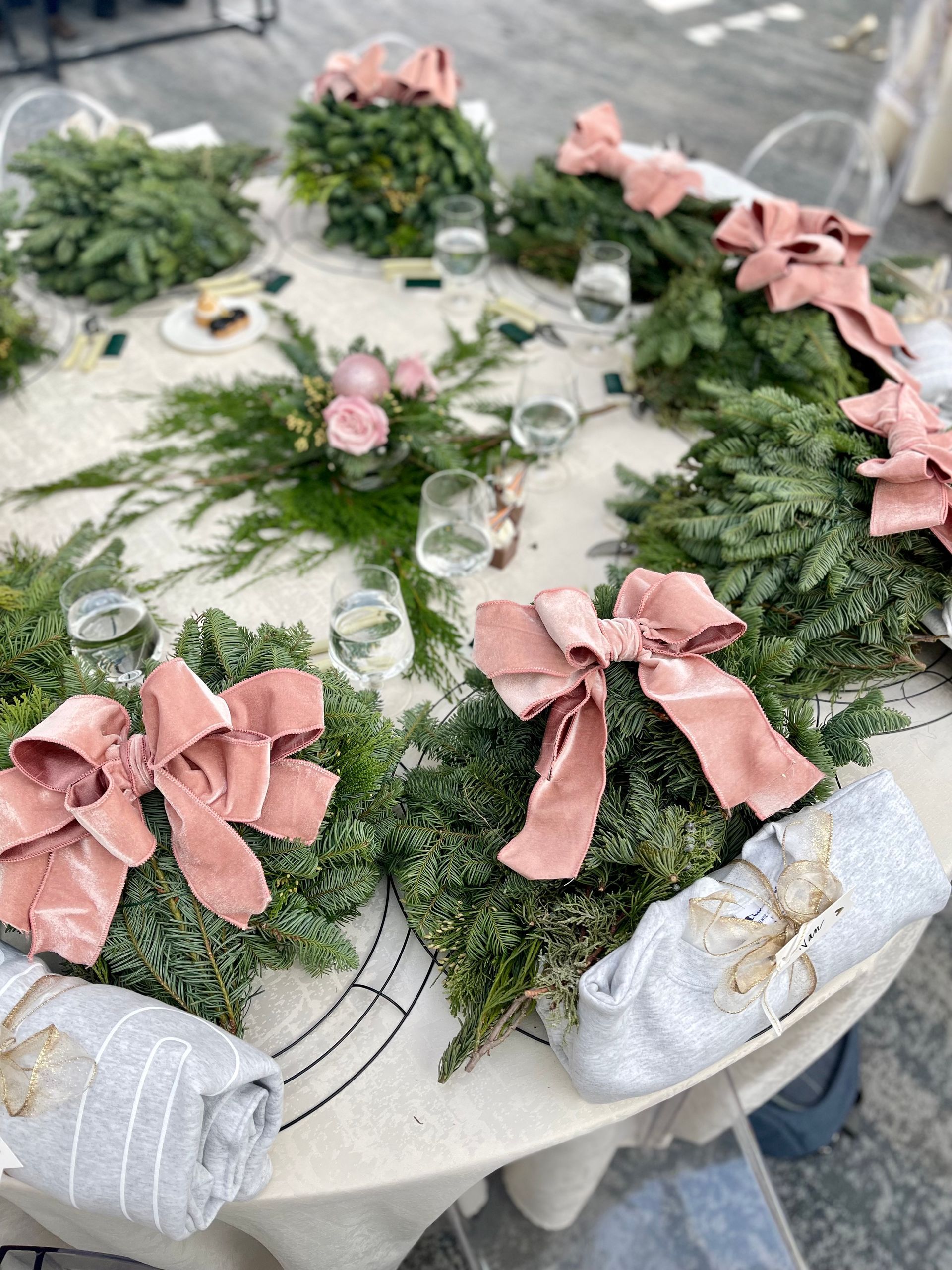 Round table set for a festive gathering, decorated with greenery, pink bows, and glassware.