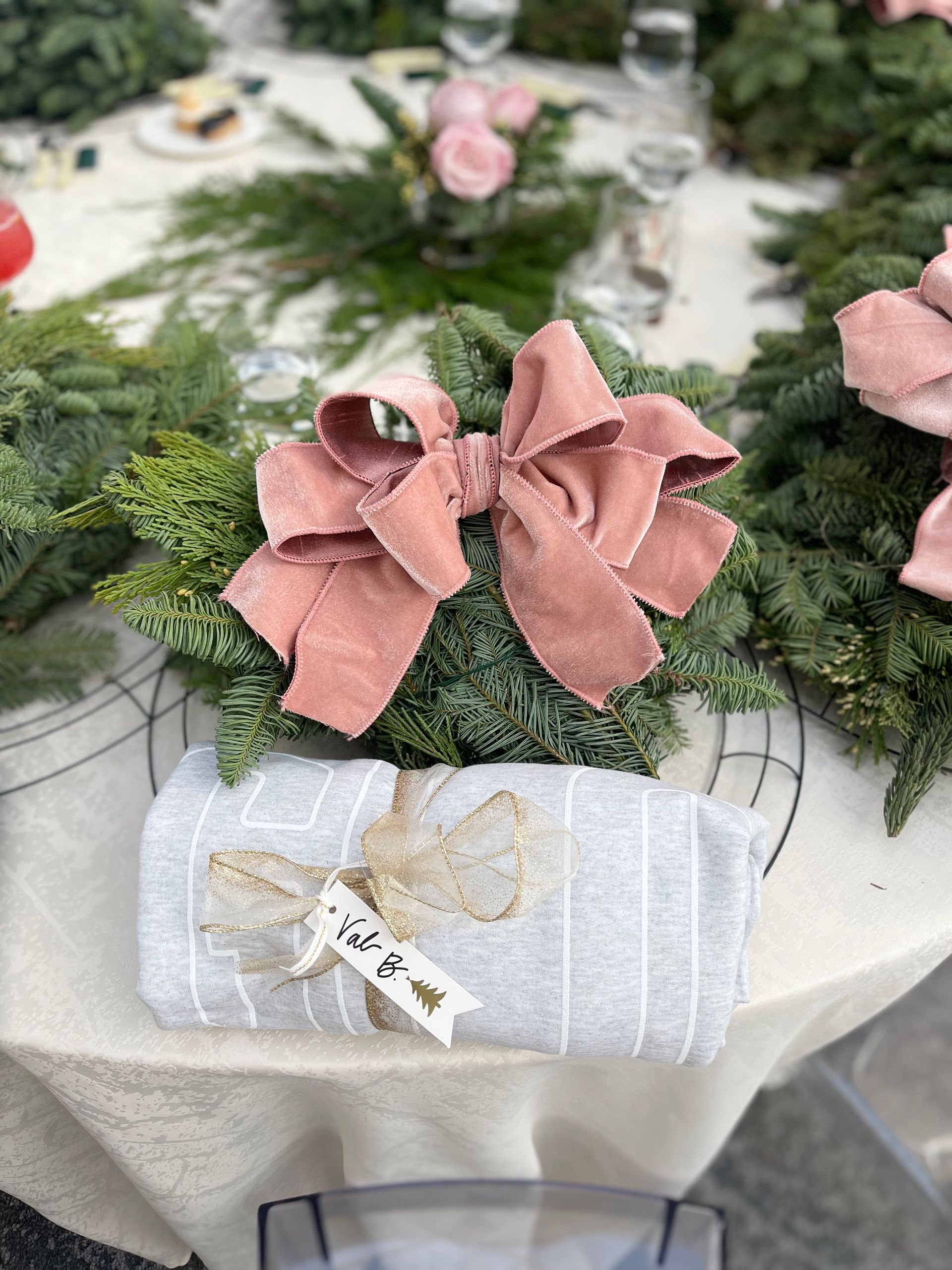 Close-up of a table setting with a pink bow on greenery, a folded towel with a tag, and surrounding evergreen wreaths.