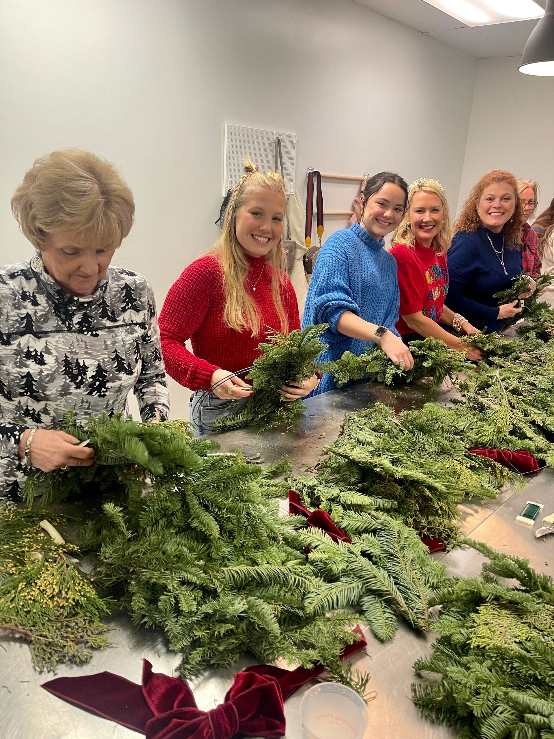 People crafting wreaths at a table. Women smiling, holding greenery. Christmas decor.