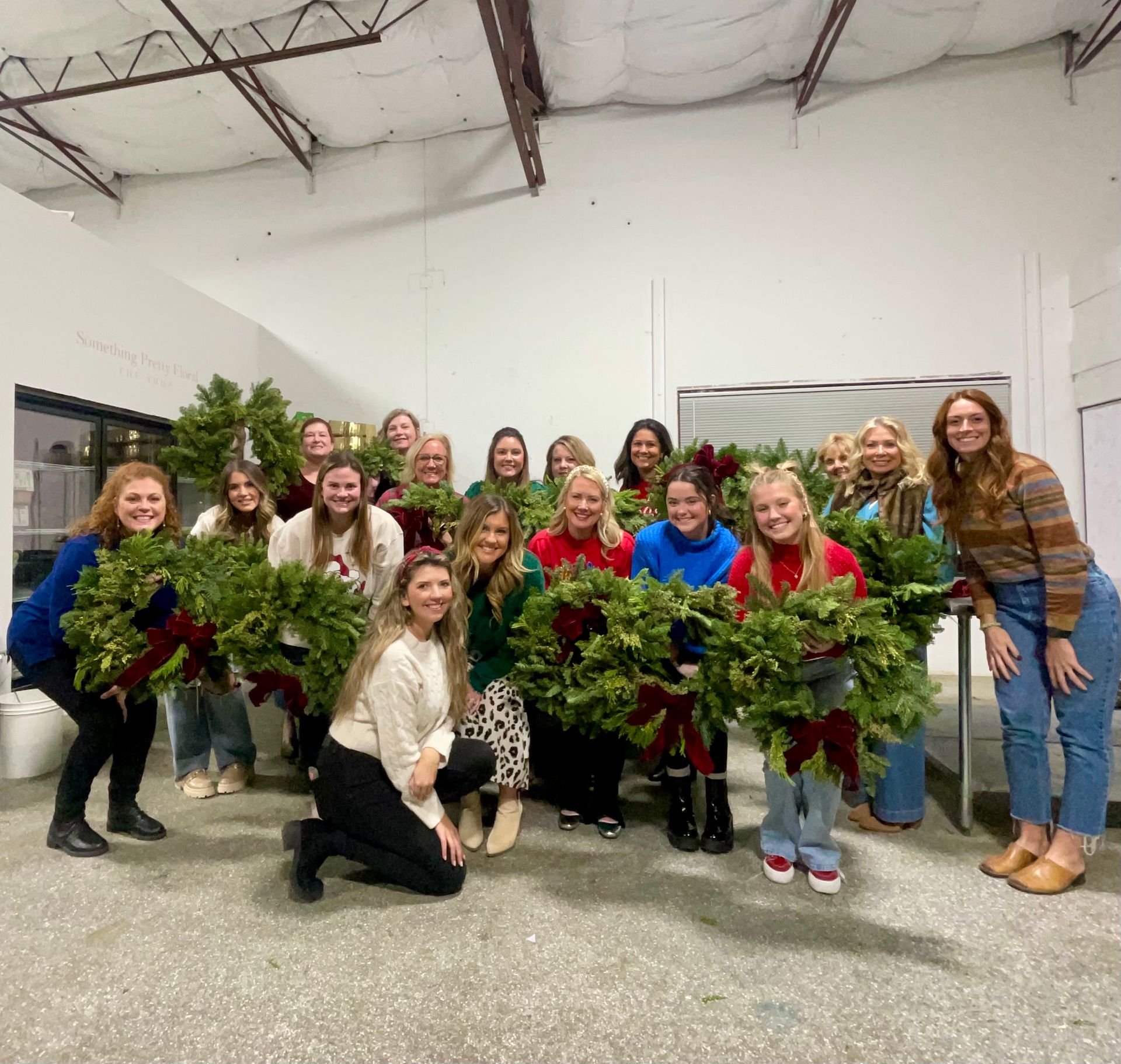 A group of women smiling, holding Christmas wreaths with red bows. Indoors, a white room.