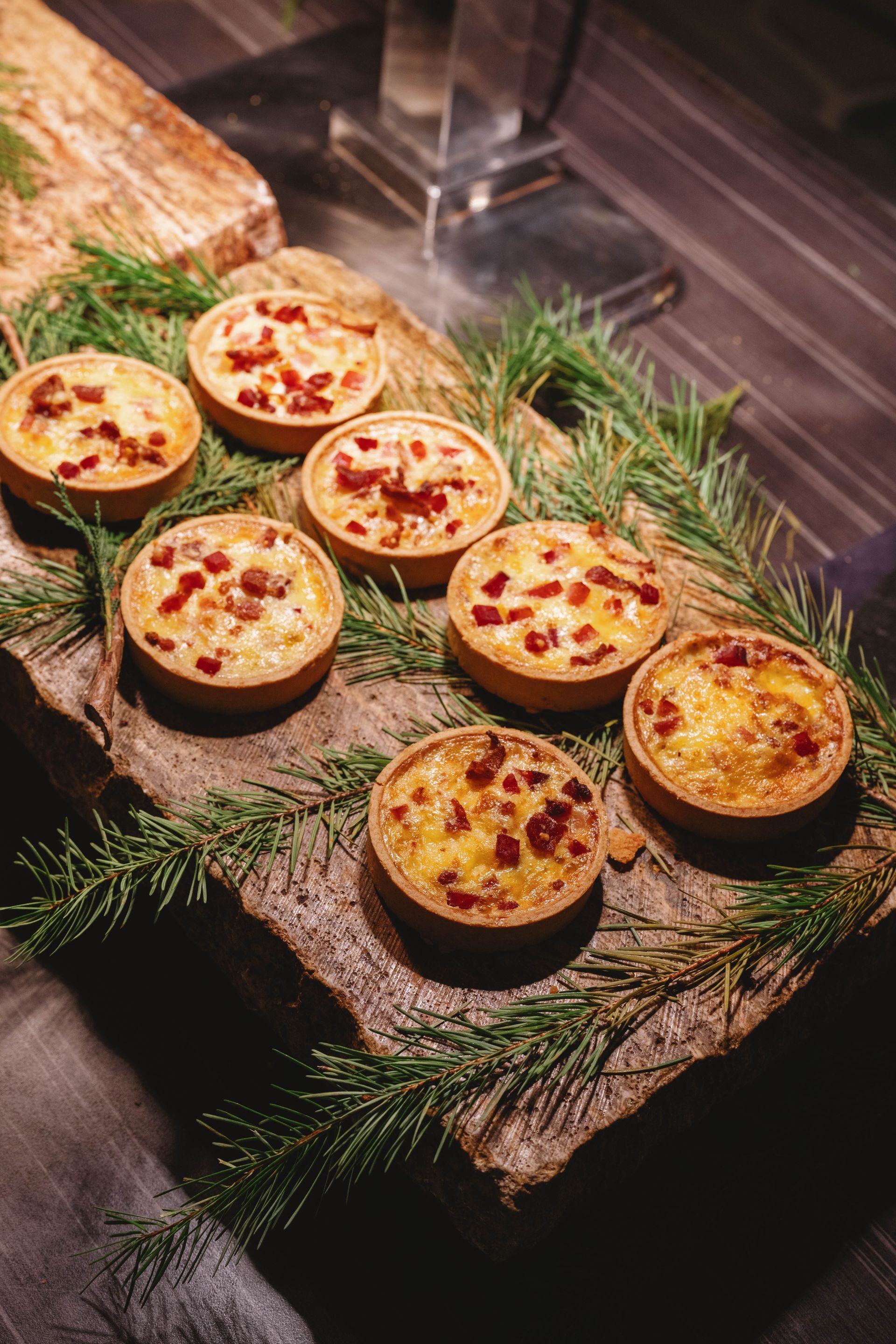 Mini quiches topped with red bacon bits on a wooden board, garnished with evergreen.
