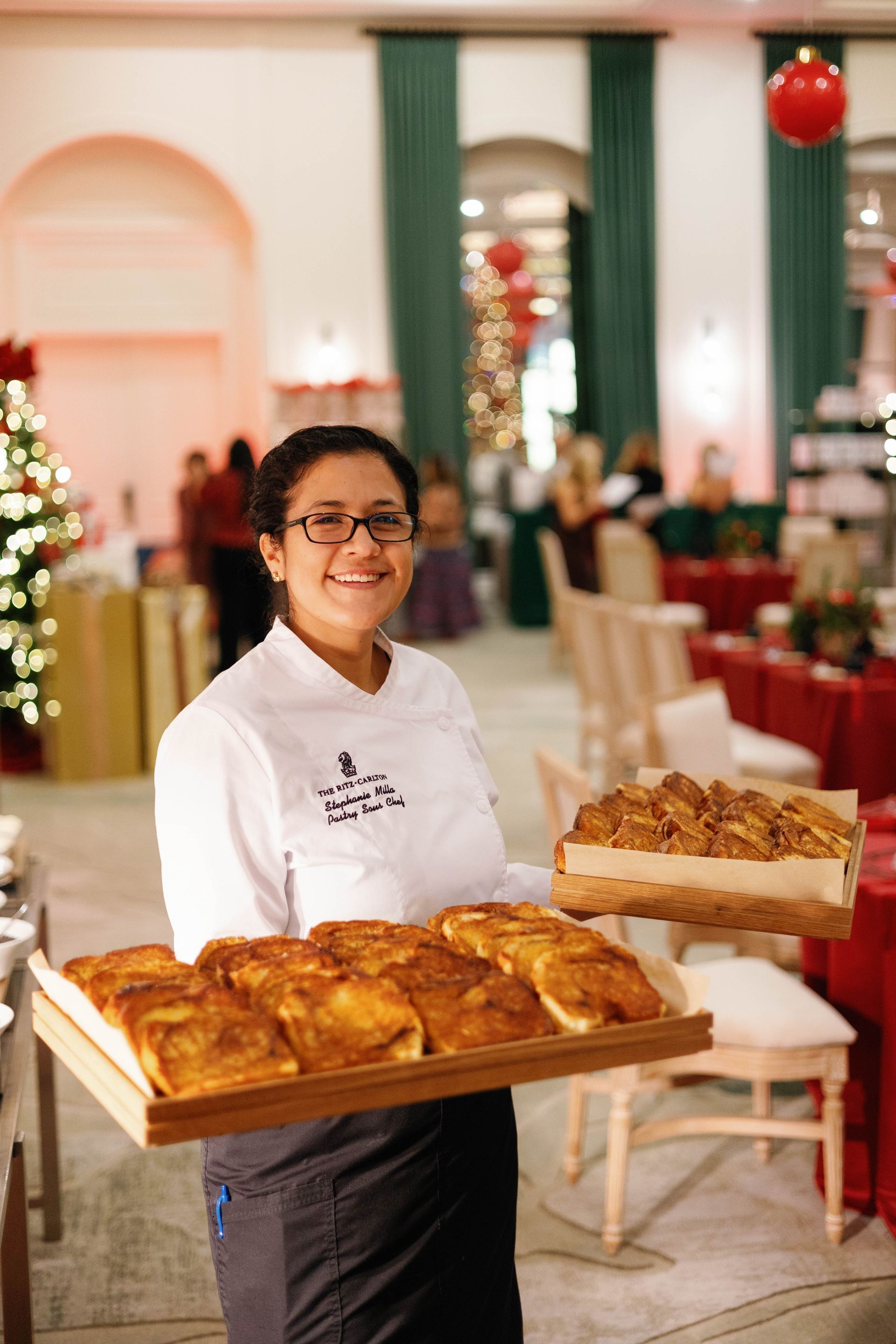 Chef holds trays of pastries, smiling in a festive dining room with holiday decorations.