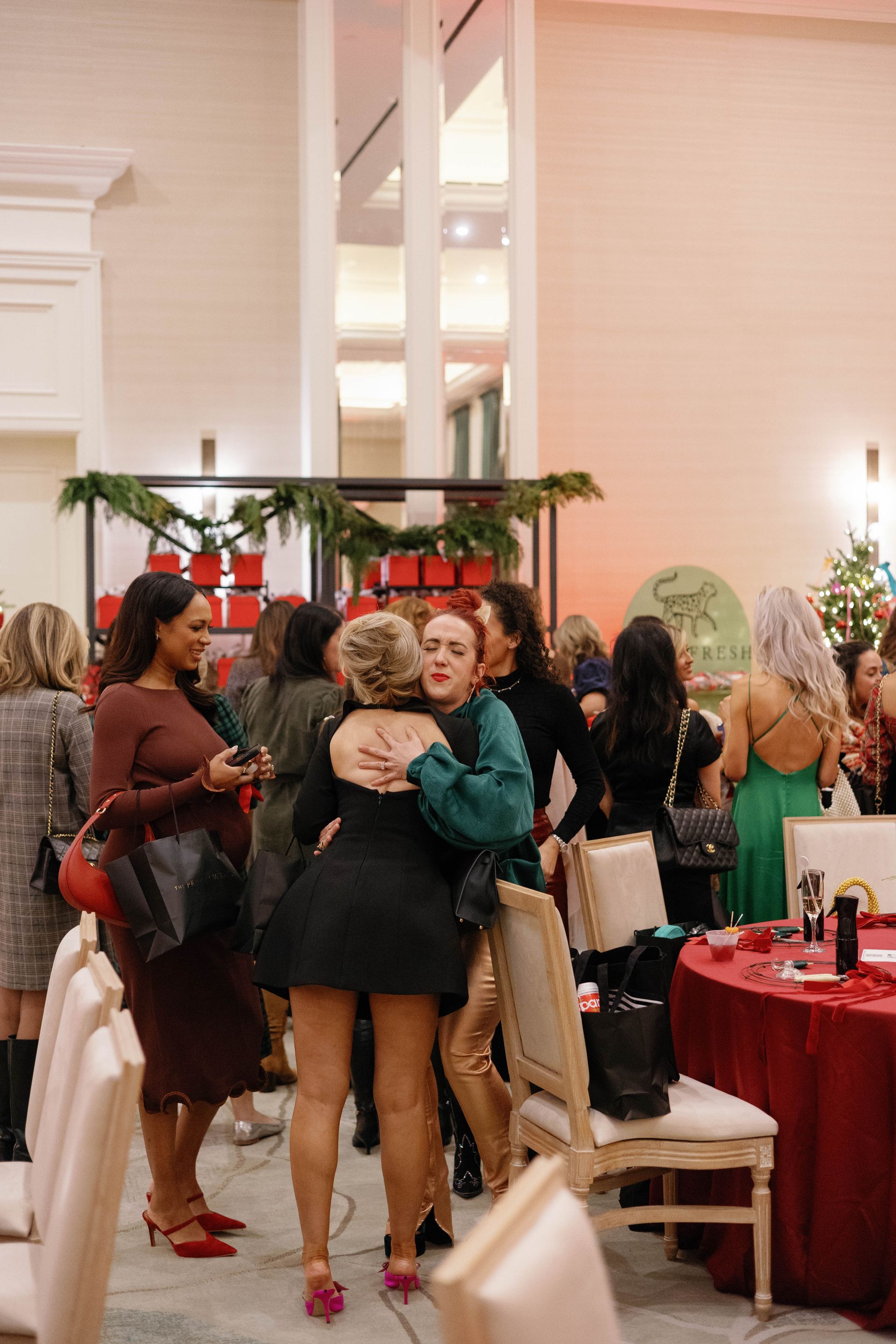 Group of women hugging at a holiday party in a decorated hall with festive decor and tables.