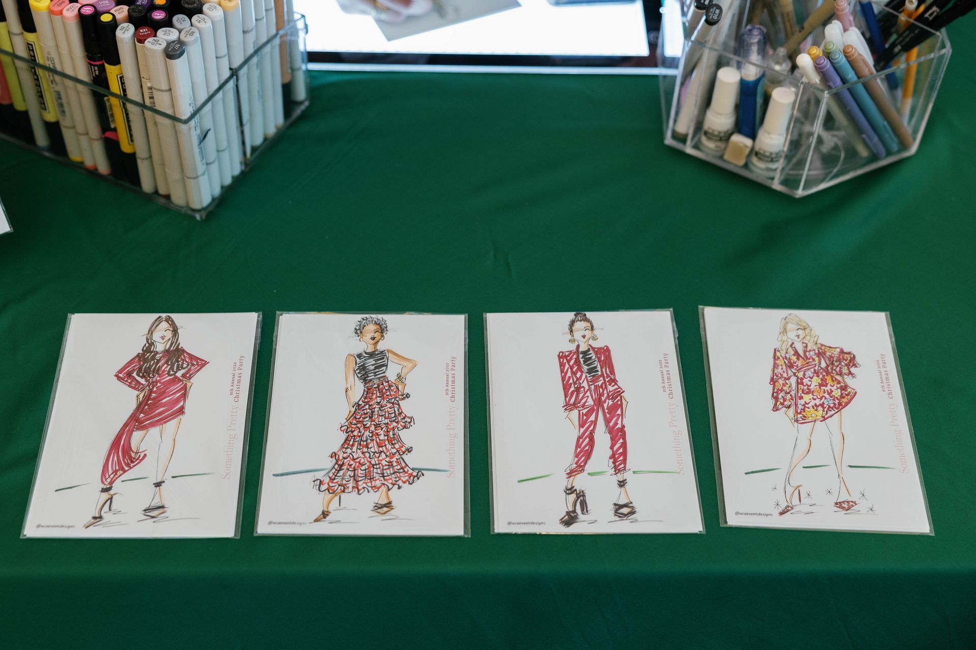 Fashion sketches on display: Four drawings of women in red and patterned outfits. Art supplies on a green table.