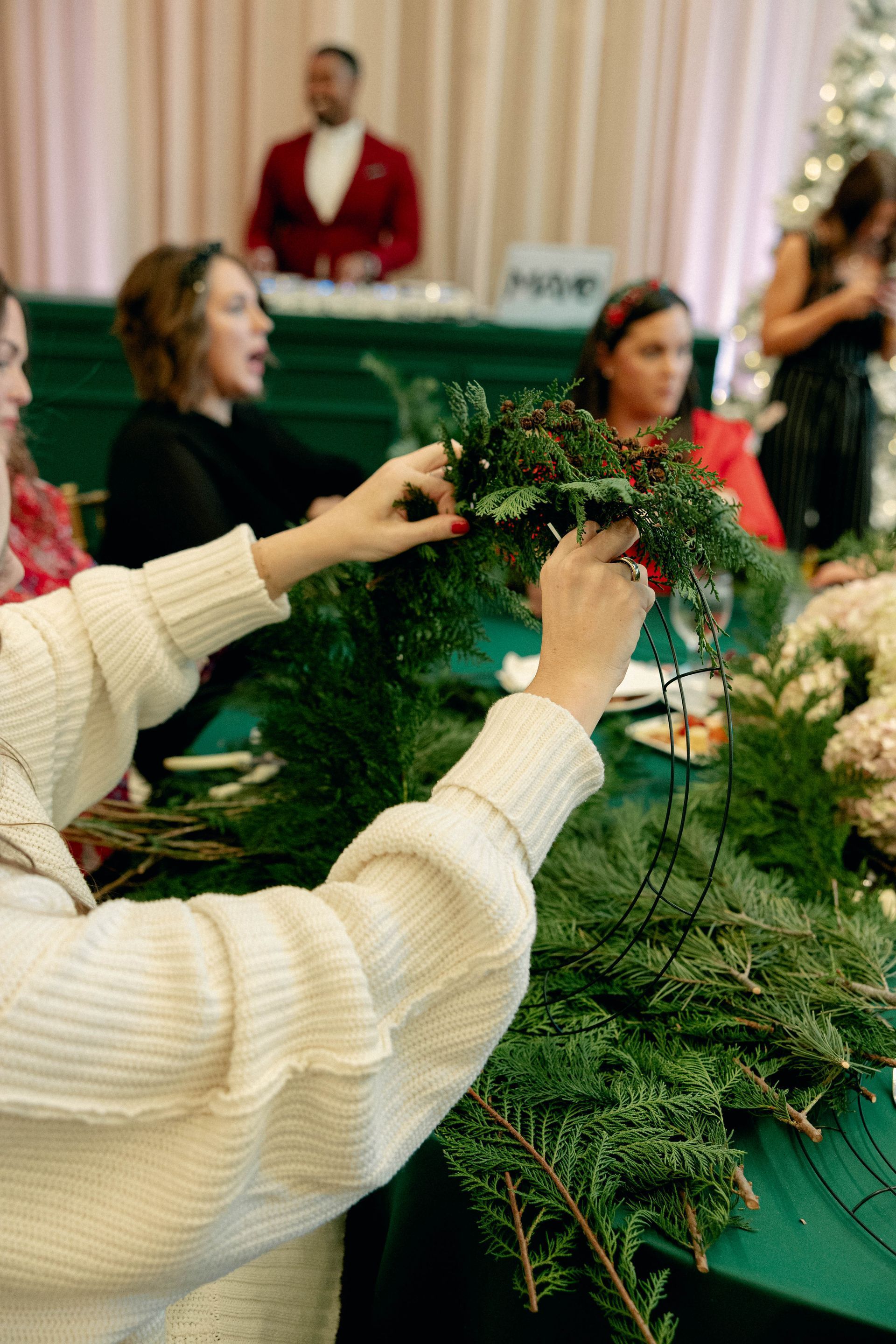 People decorating wreaths at a table. Woman in a white sweater adds greenery. Others look on. Festive setting.