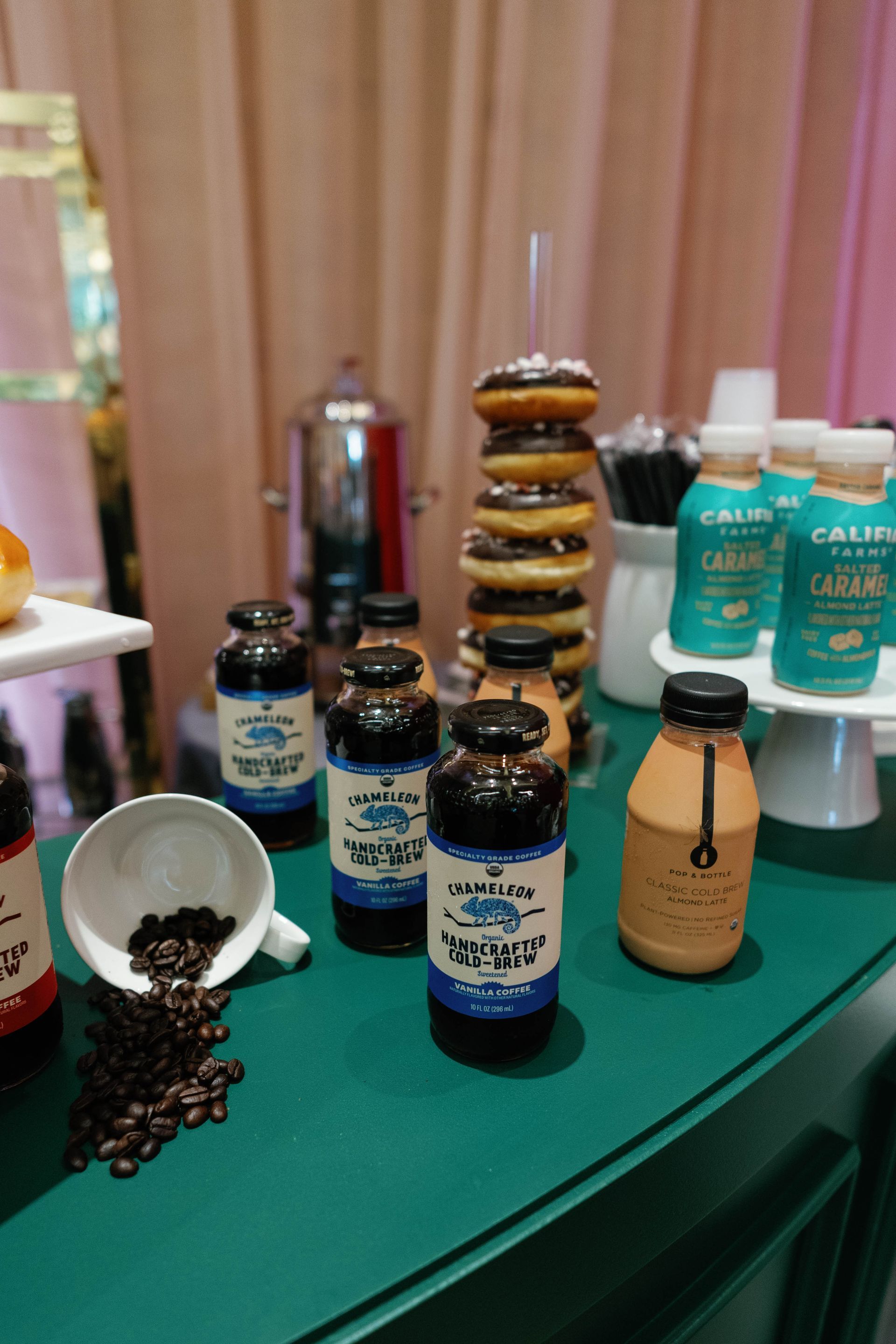 Coffee bar: bottles, donuts, coffee beans, and drinks arranged on a green table, pink backdrop.