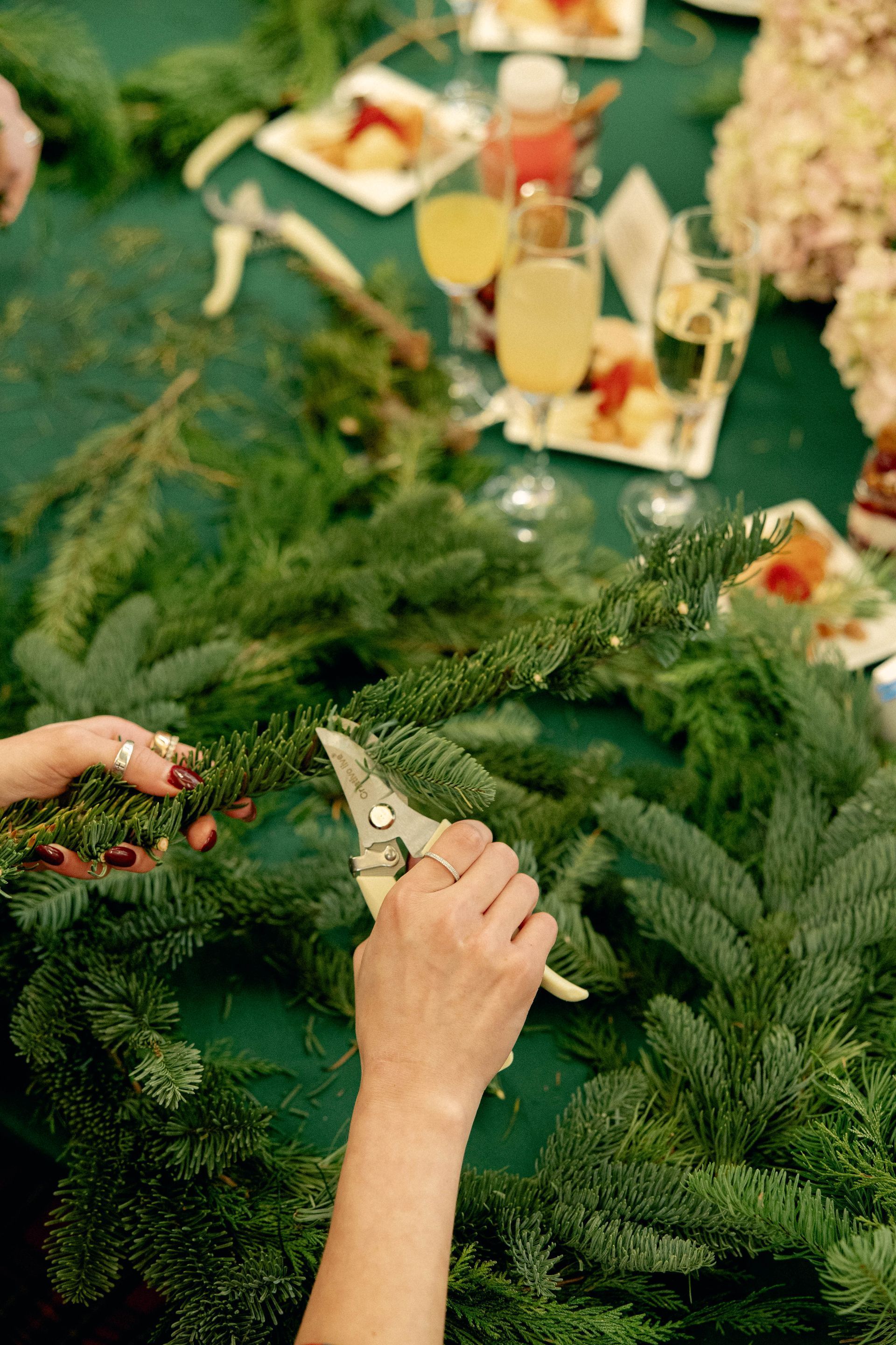Woman's hands making a wreath with shears and greenery, drinks and food on a green table.