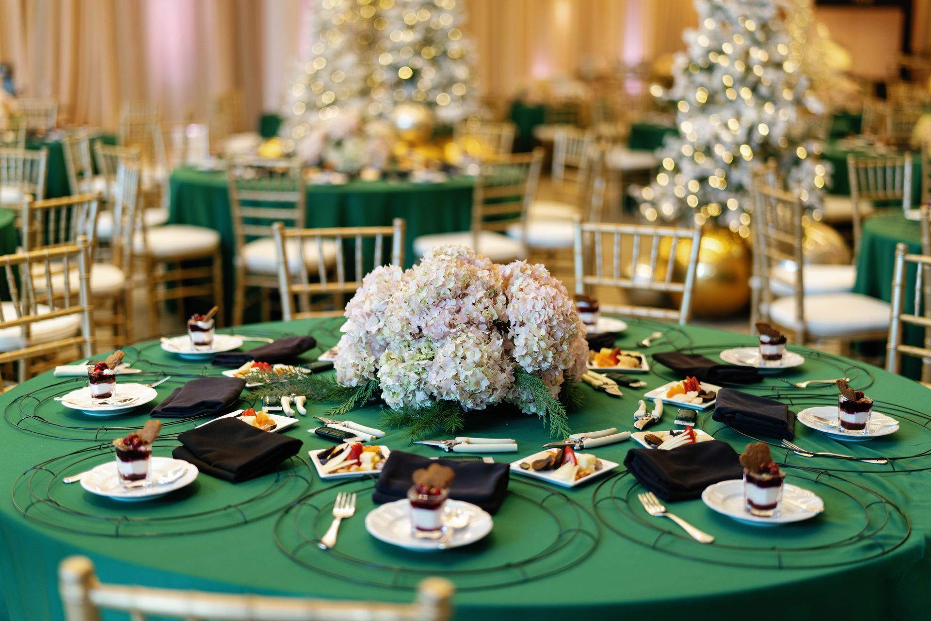 Elegant dining room with green tablecloths, gold chairs, and Christmas trees; table settings with food and flowers.