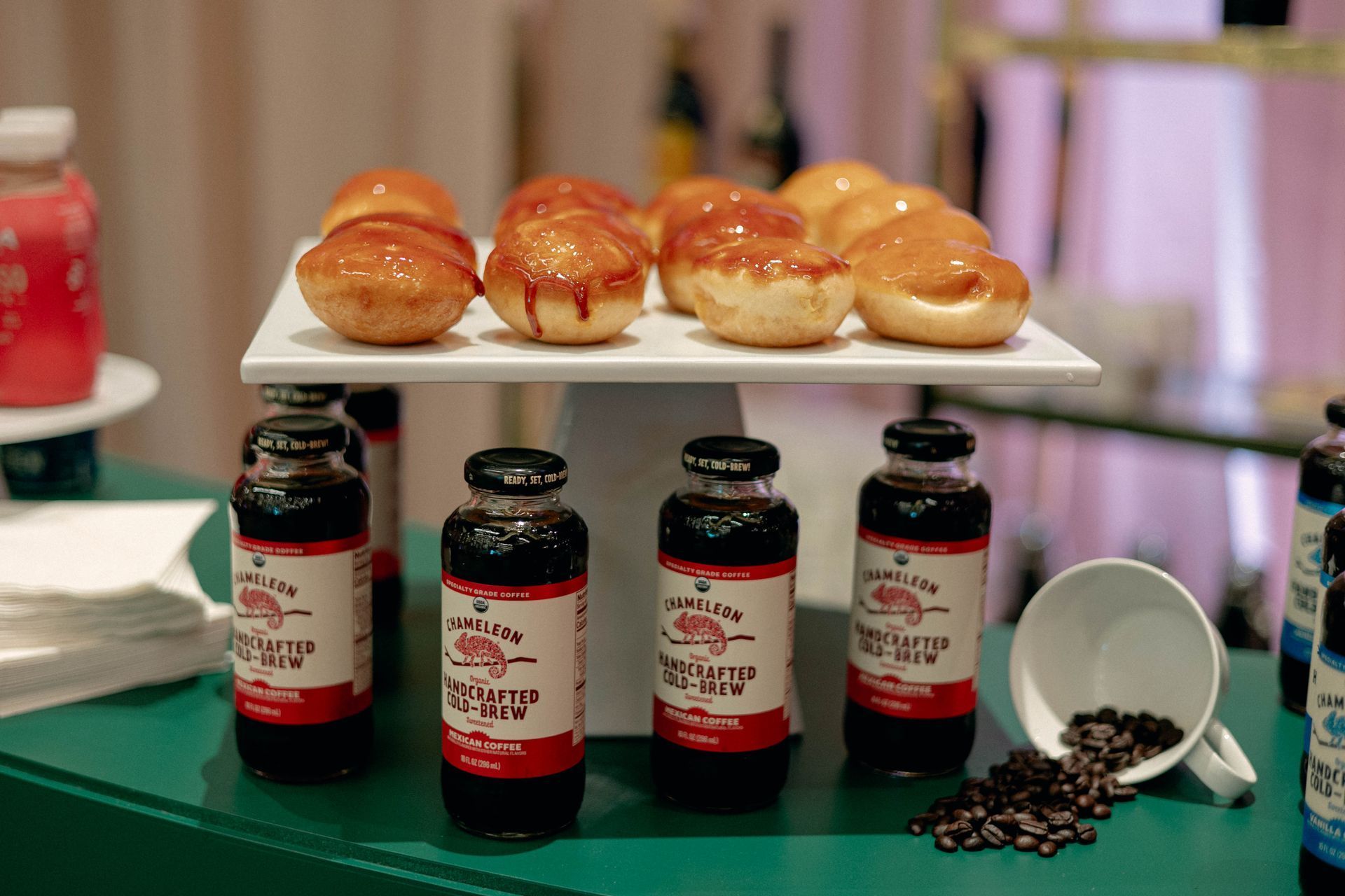 Bottled iced coffee and glazed donut pastries on a white stand, on a green surface. Coffee beans spill from a mug.