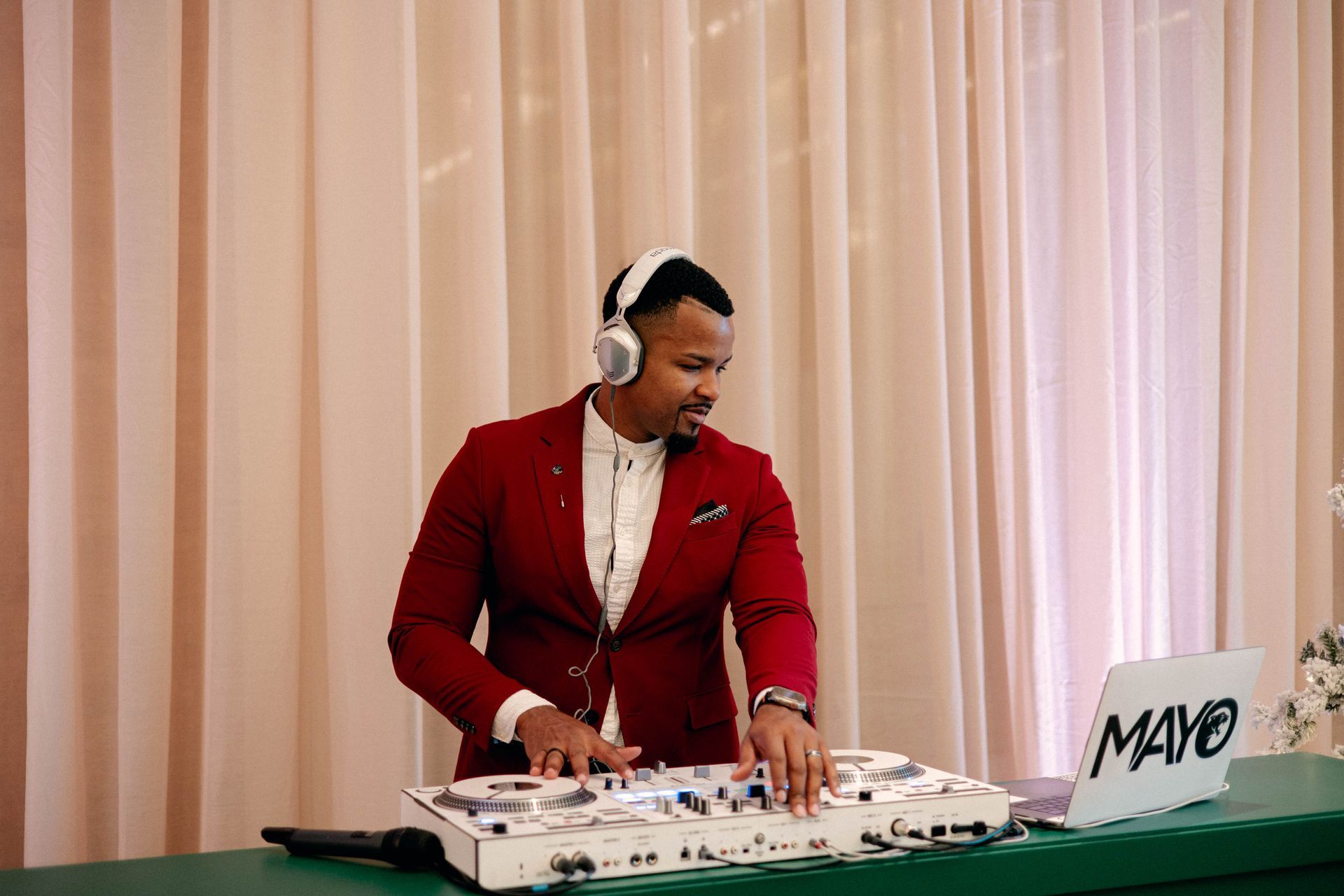 DJ in a red suit, wearing headphones, mixing music at a wedding reception with a laptop labeled 