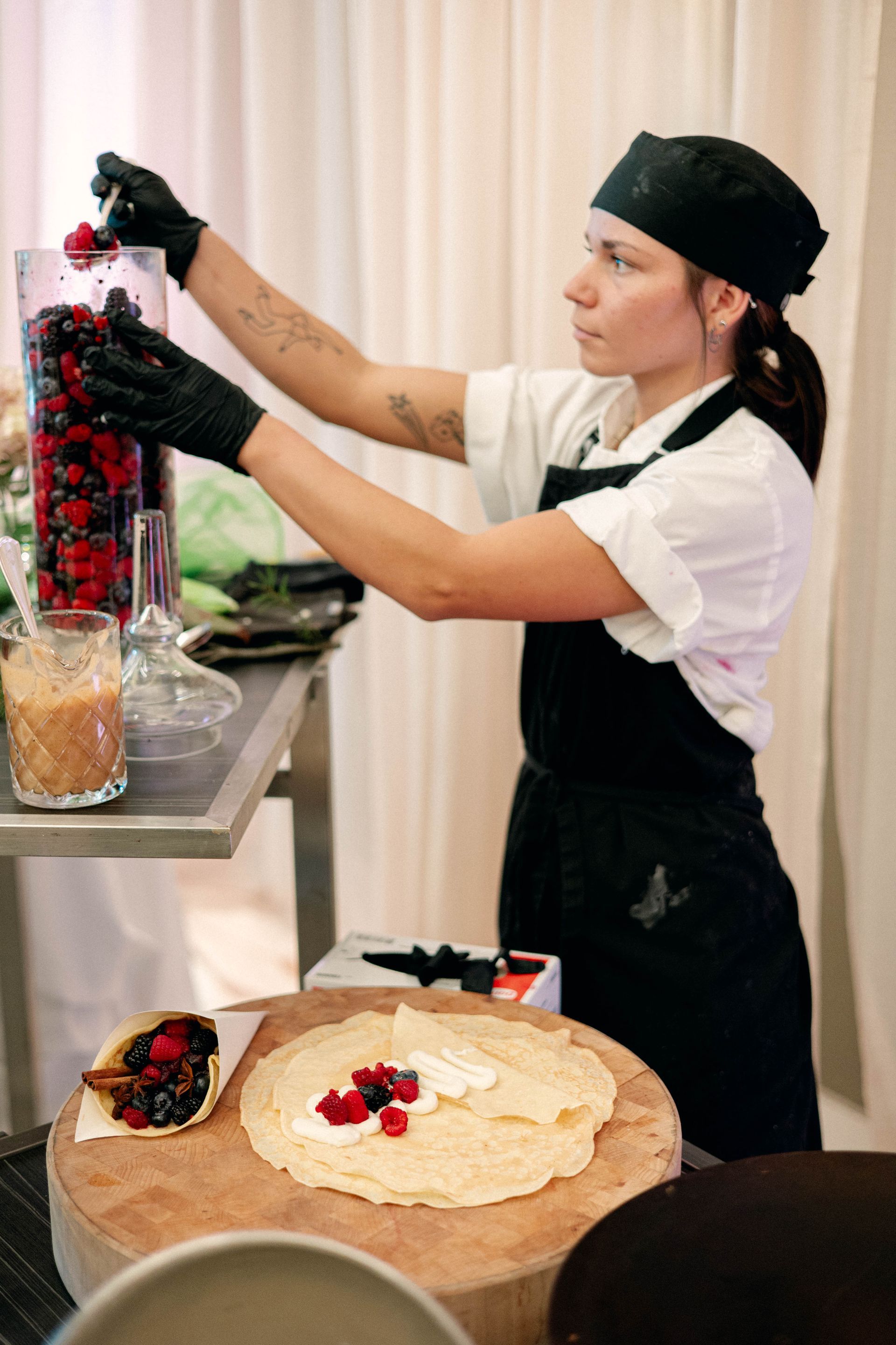 Woman in chef's attire adding berries to crepes on a wooden board.