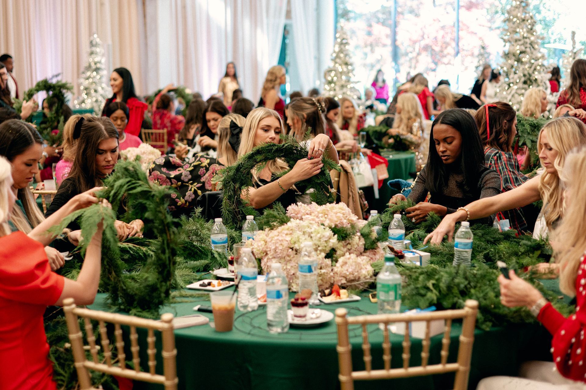 Women at a table crafting holiday wreaths. Green tablecloth, gold chairs, Christmas trees in background.