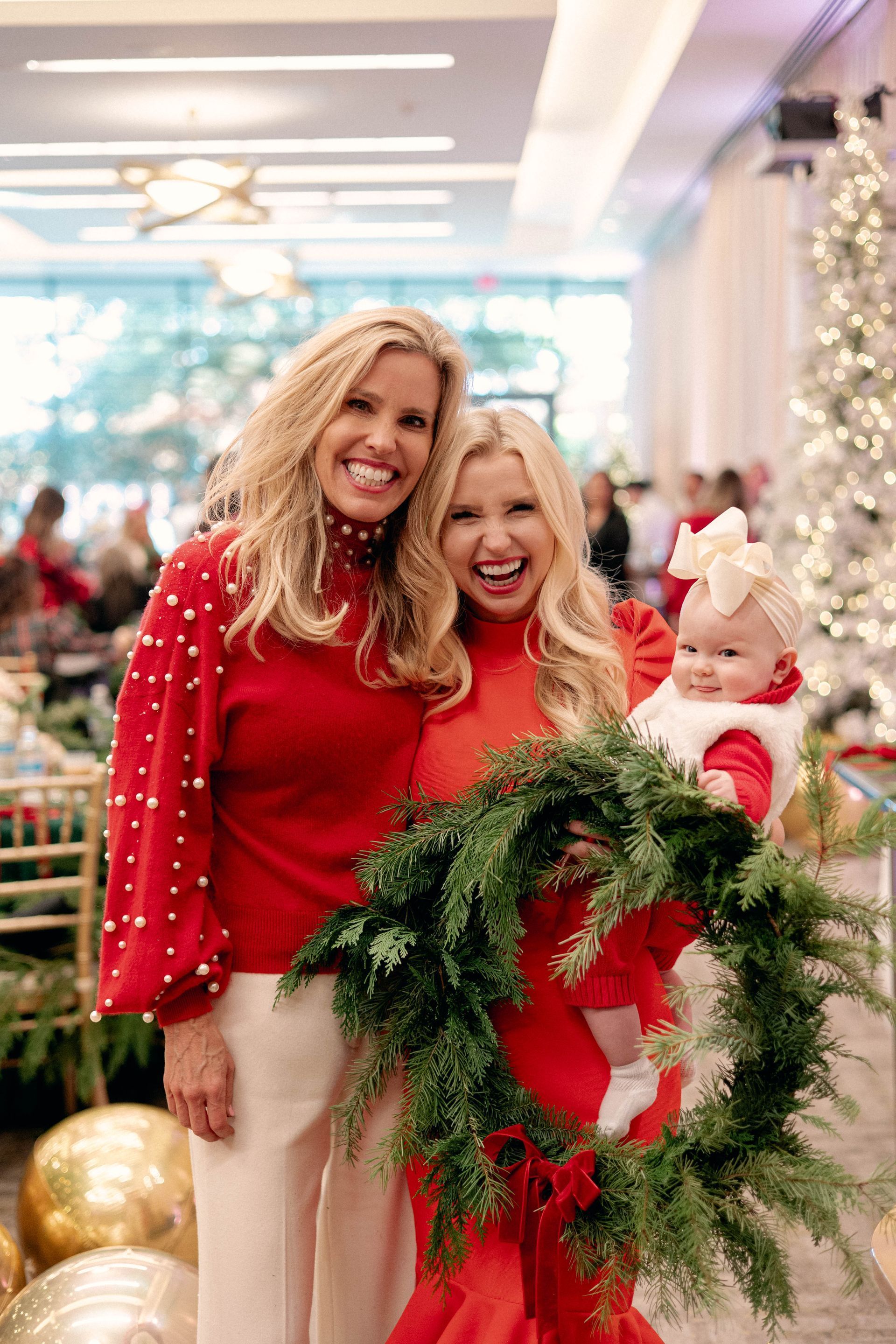 Three women in red outfits pose with a baby. One holds a wreath in a festive setting.