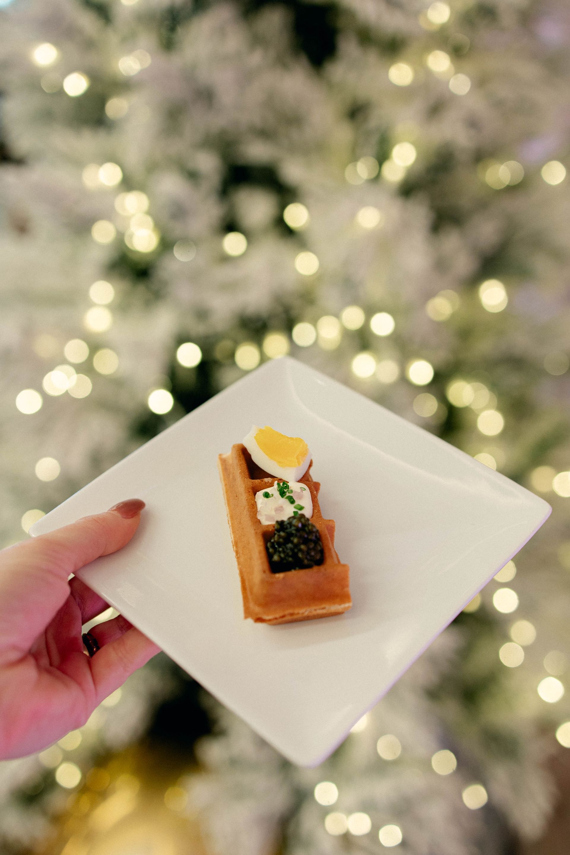 Hand holding a plate with a gourmet appetizer: waffle topped with caviar, egg, and cream, festive lights in background.