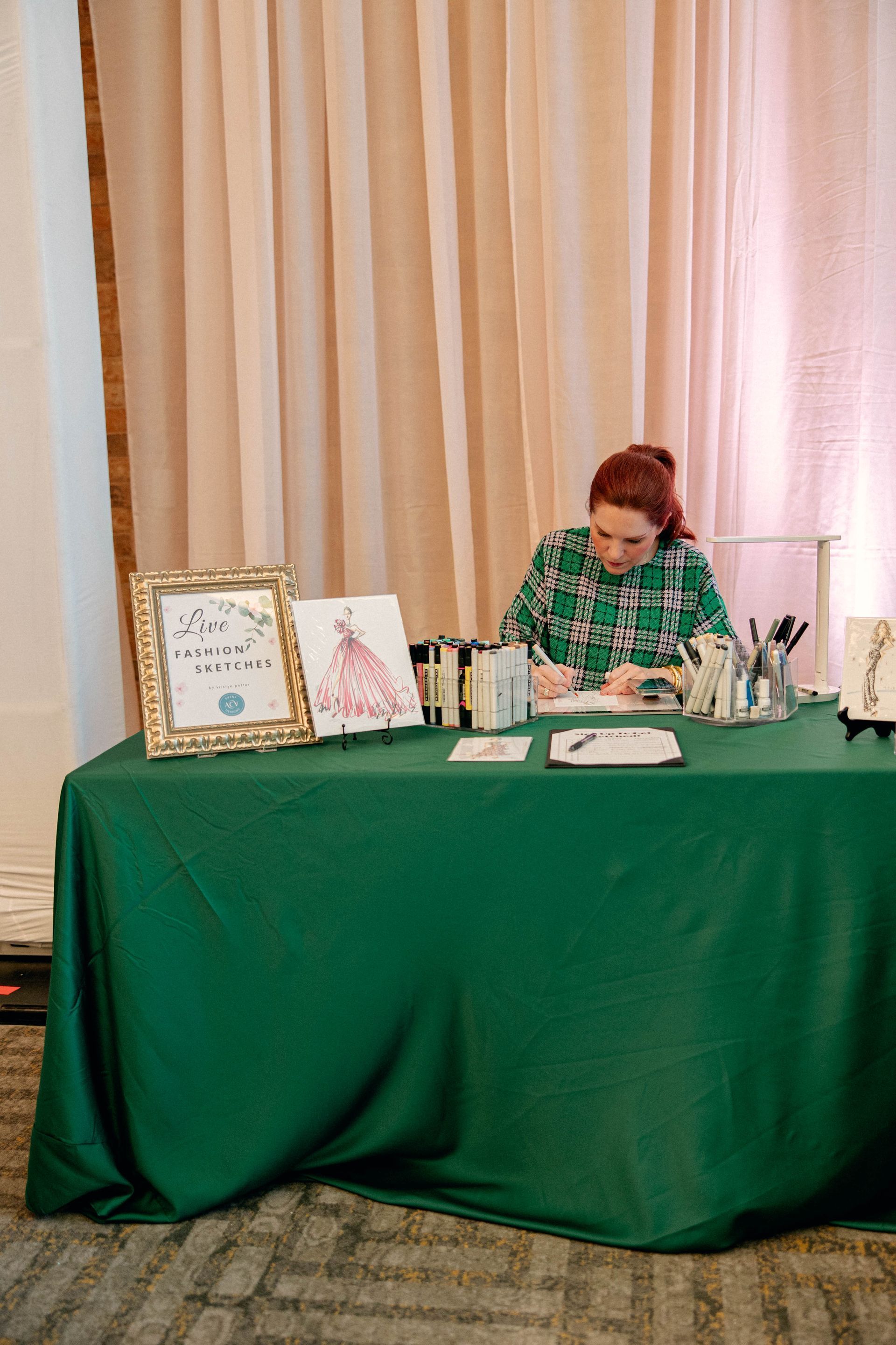 Woman sketching at a green-covered table, with framed art, supplies, and pink curtains behind her.