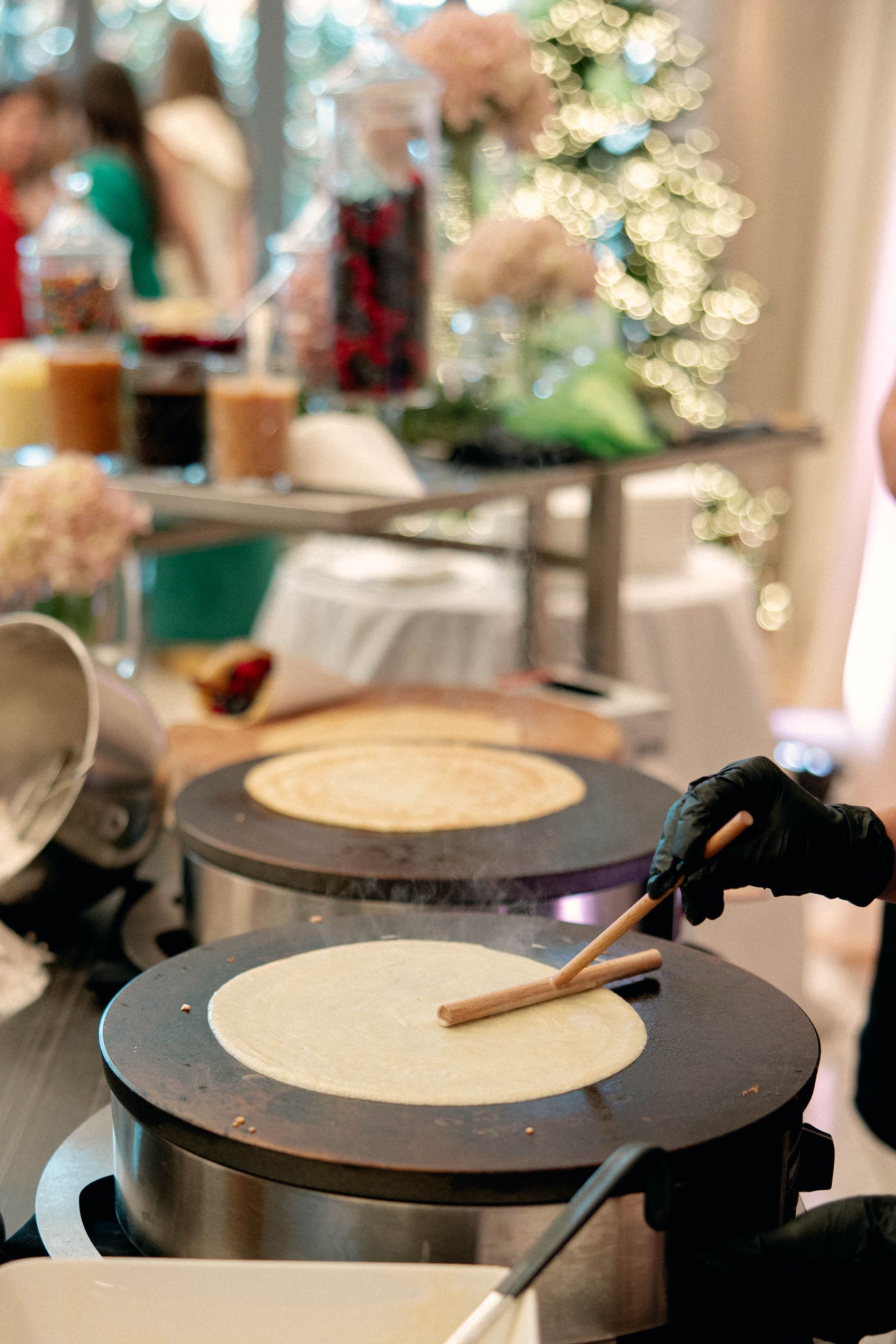 Crepe station with two griddles, person in gloves cooking crepes, toppings buffet in background.