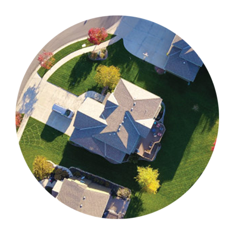 Aerial view of suburban houses with roofs, lawns, trees, and a curved driveway in a neighborhood.