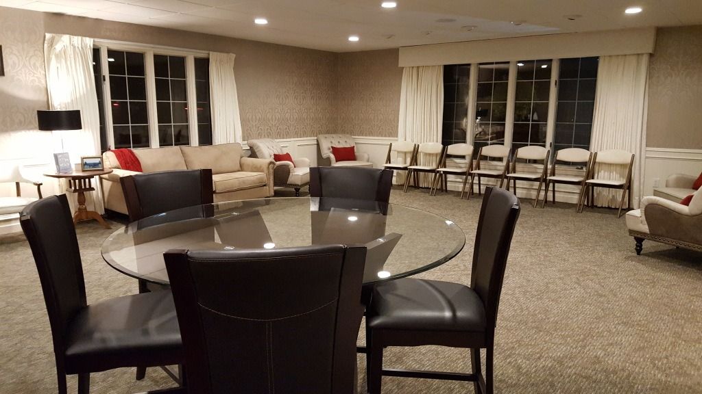 A room with a round table and chairs, a couch, windows with curtains, and folding chairs.