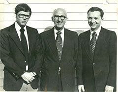 Three men in suits stand together, smiling.