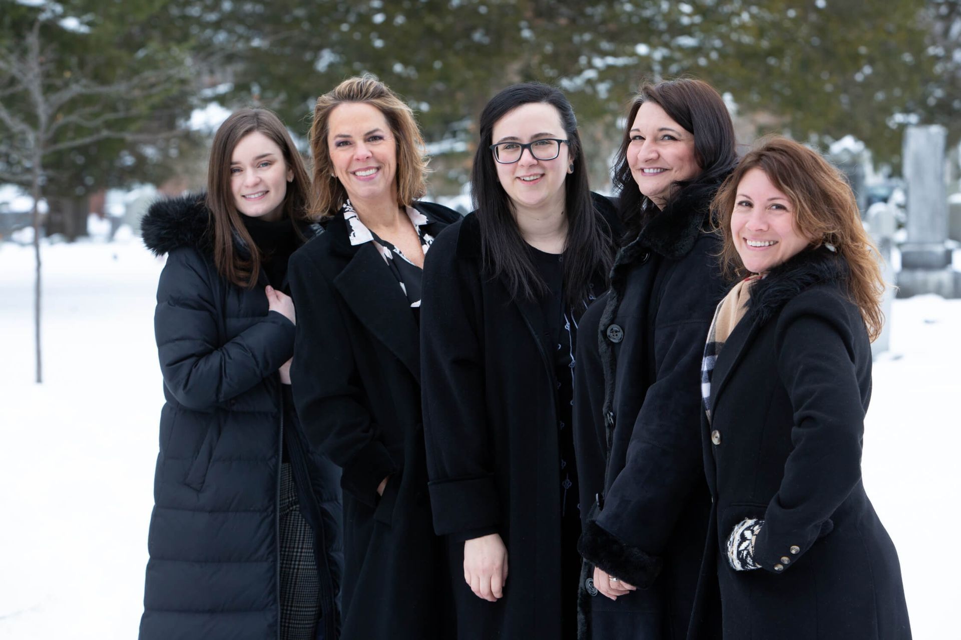 Five women in black coats pose in a snowy cemetery.