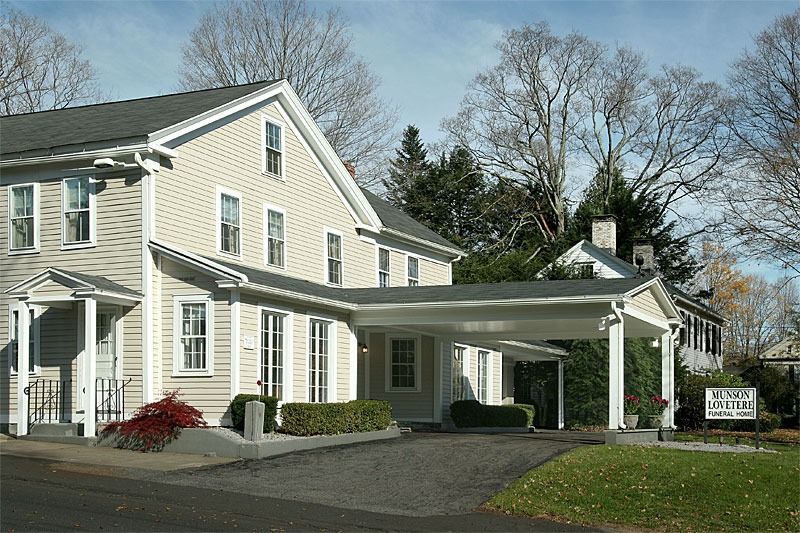 Cream-colored building with portico, likely a funeral home, surrounded by trees and lawn.