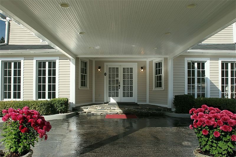 Covered entrance of a light-colored building with glass doors, framed by windows, flanked by red flowers.