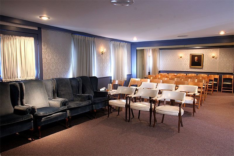 Interior of a funeral home chapel with rows of chairs and blue seating. Light-colored walls and carpet.
