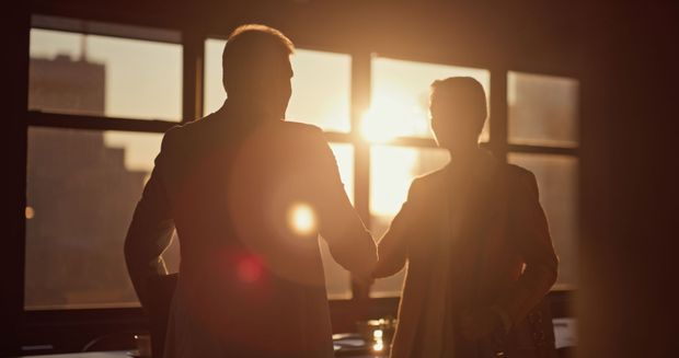 A man and a woman are shaking hands in front of a window at sunset.