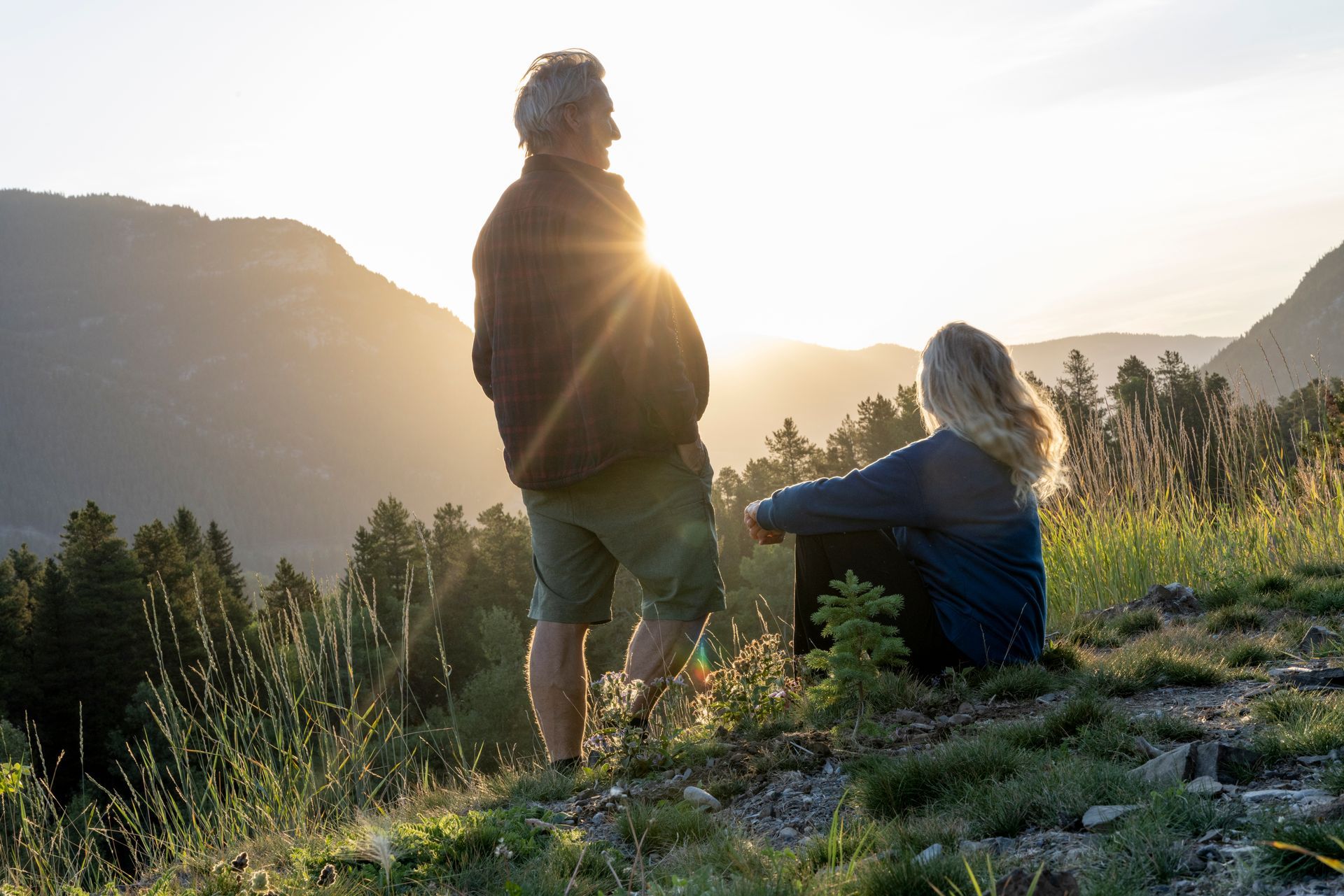 A man and a woman are standing on top of a hill looking at the sunset.