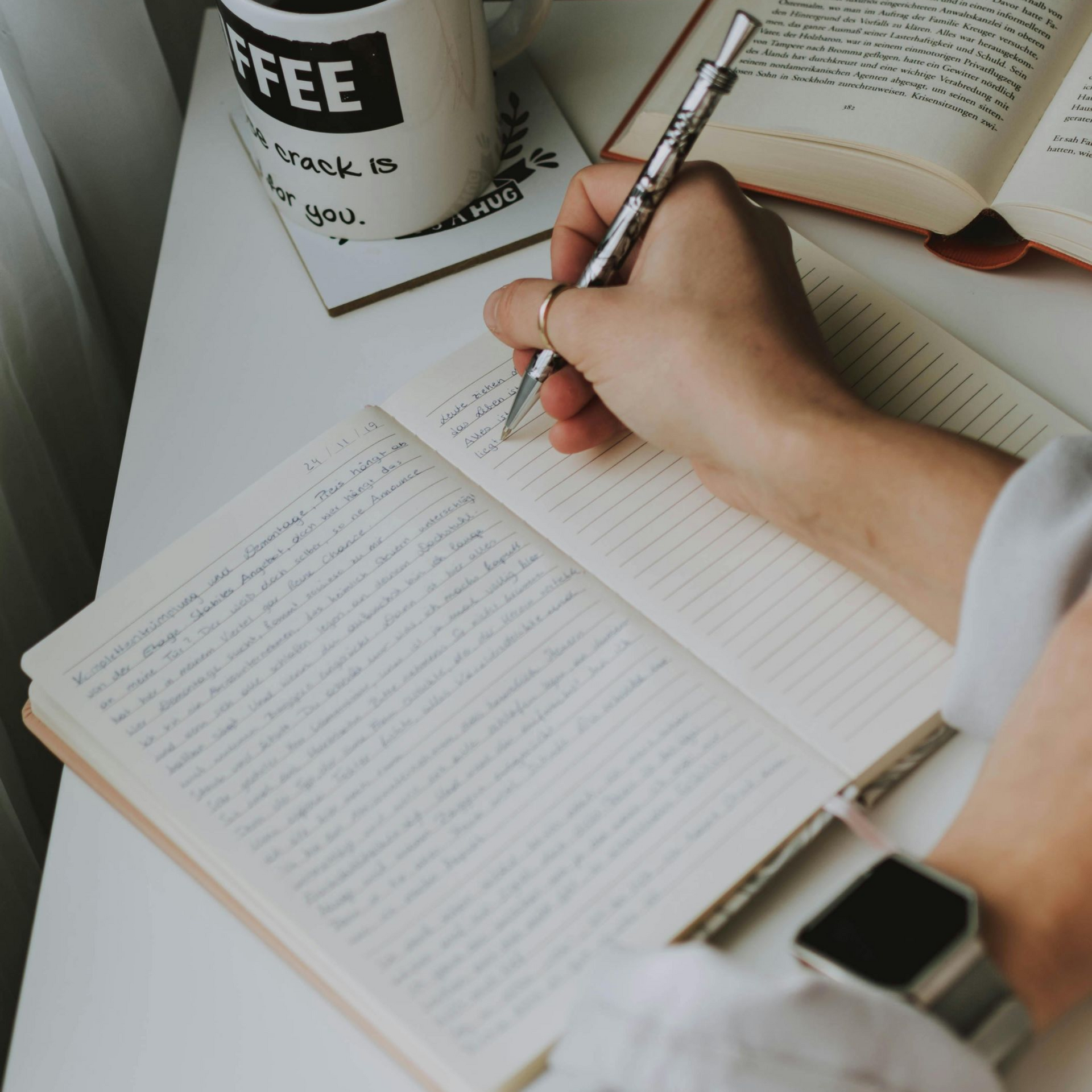 Person writing in a notebook, next to a coffee mug and open book on a white desk.