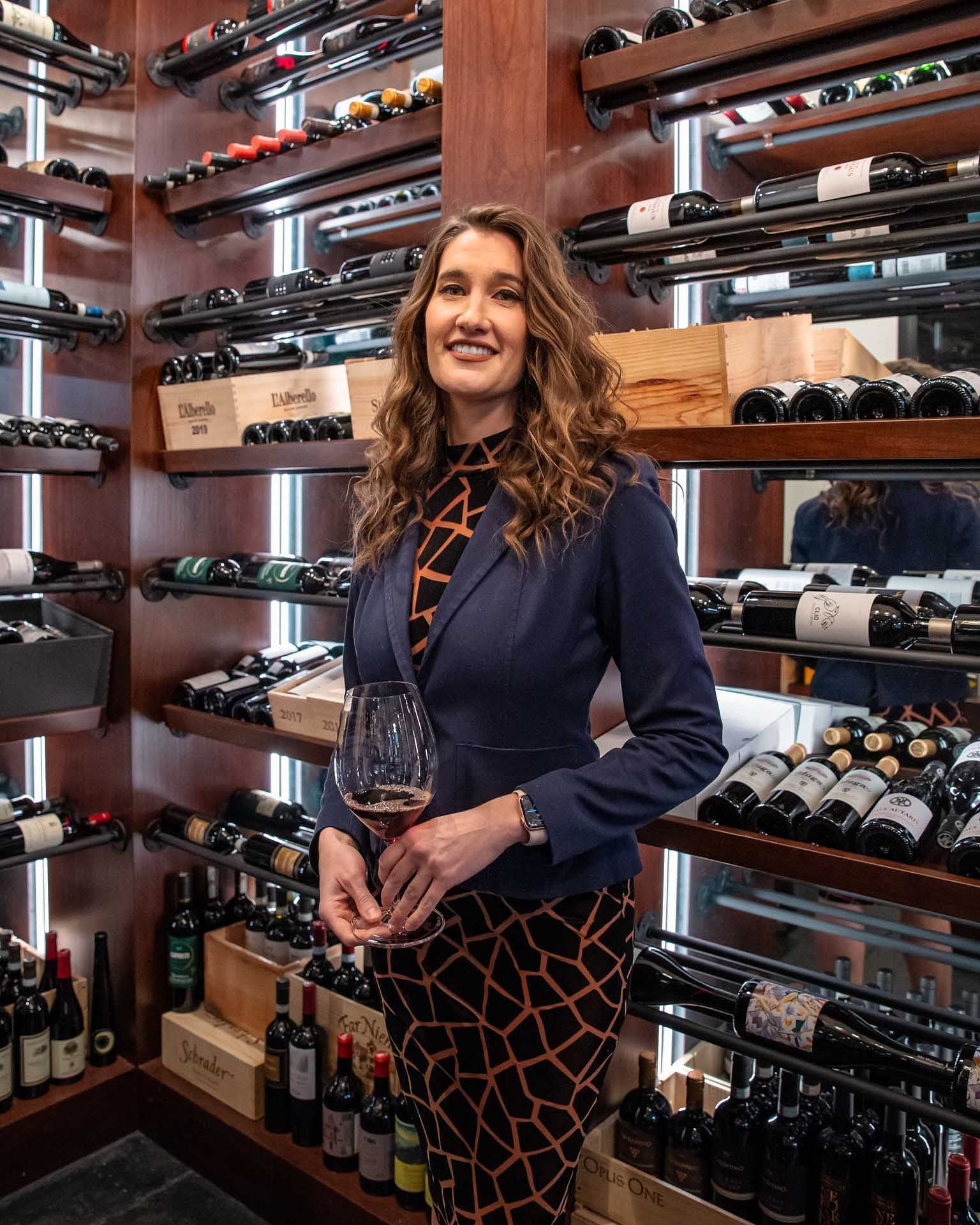 bethany morris holding a glass of wine in front of a wall of wine bottles including a bottle of lagrein