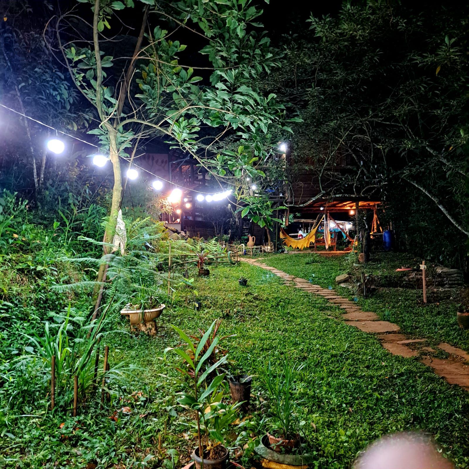 Cena noturna com um caminho de jardim iluminado que leva a uma estrutura. Luzes de cordão iluminam o espaço; vegetação exuberante circunda a paisagem.