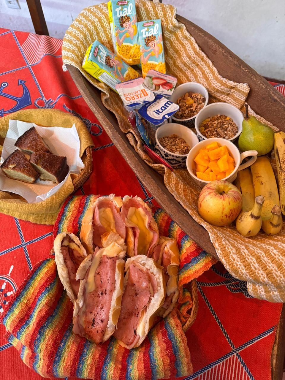 Café da manhã variado: cereais diversos, iogurte, frutas, torradas e bacon em cestas sobre uma toalha de mesa estampada.