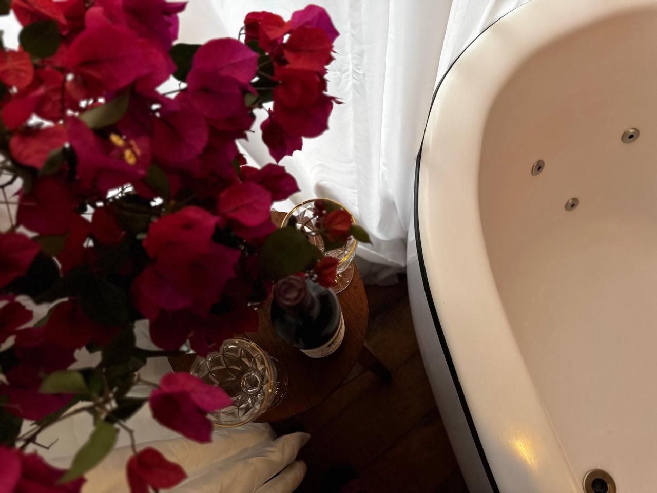 Flores cor-de-rosa ao lado de uma banheira branca, com vinho e taças sobre uma pequena mesa.