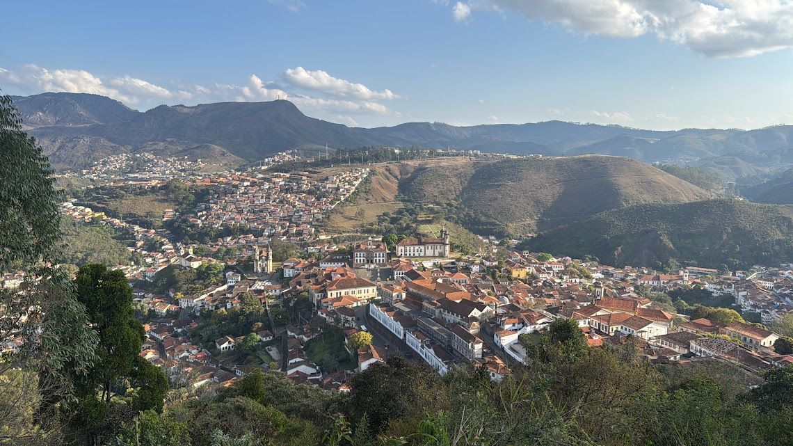 Vista panorâmica de uma cidade aninhada em um vale, rodeada por colinas e montanhas verdejantes sob um céu azul.