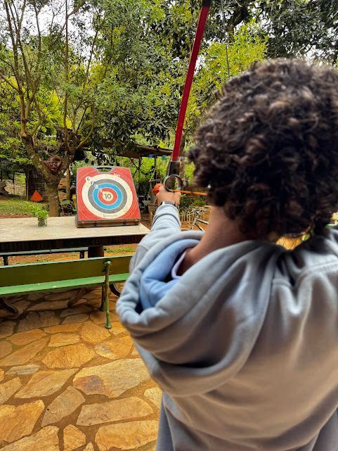 Pessoa mirando uma flecha em um alvo ao ar livre. Arco vermelho, moletom azul, alvo colorido.