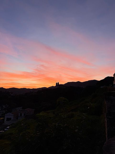 Pôr do sol sobre uma paisagem urbana; céu alaranjado e rosado acima das silhuetas escuras de prédios e colinas.