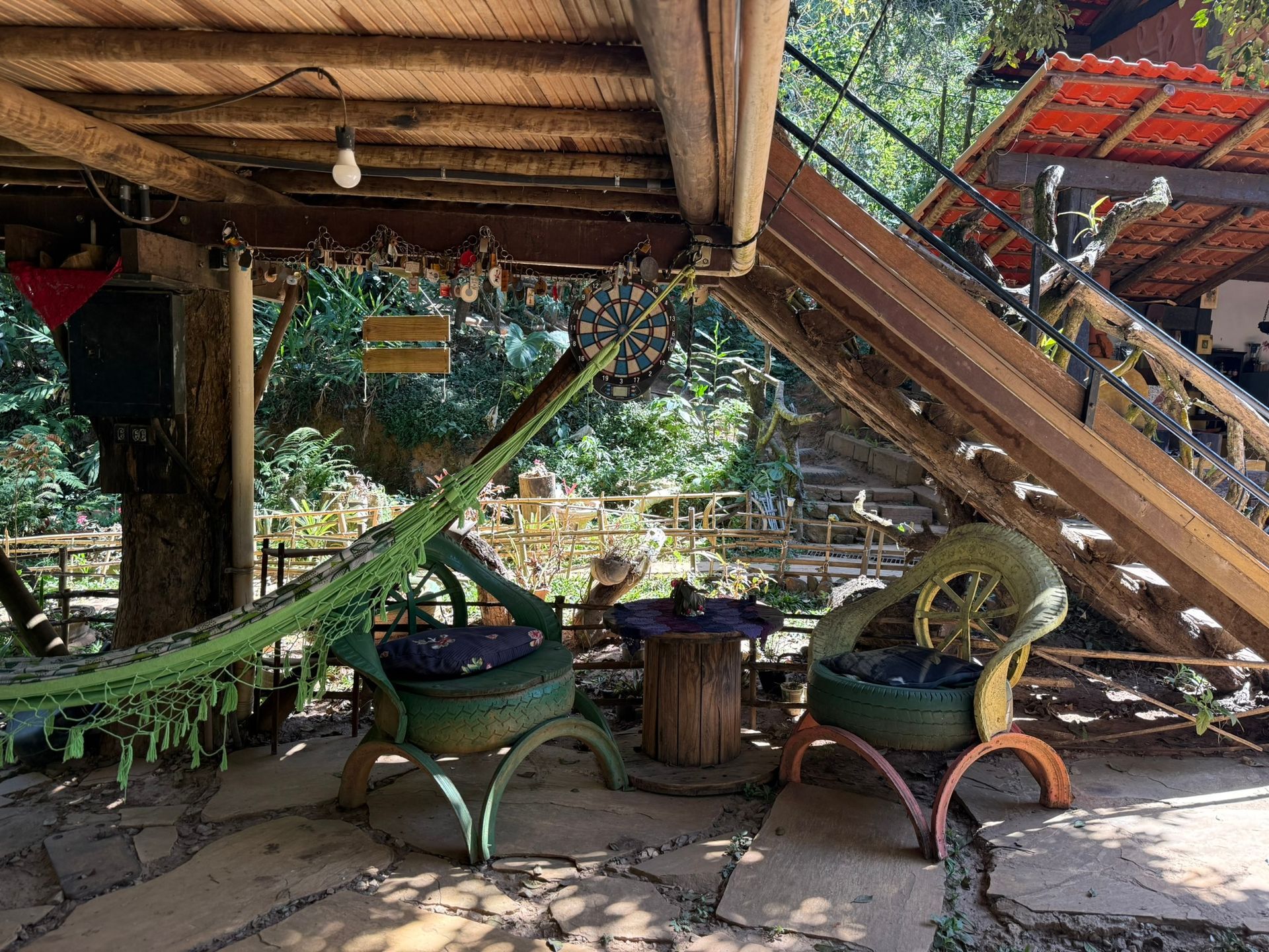 Área de estar externa com cadeiras feitas de pneus, rede, mesa de madeira, escadas e teto de palha.