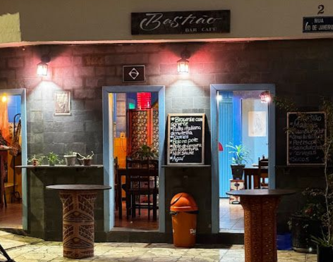 Fachada de um bar à noite, com portas azuis, luzes e mesas do lado de fora. Um cardápio escrito em um quadro-negro é visível.