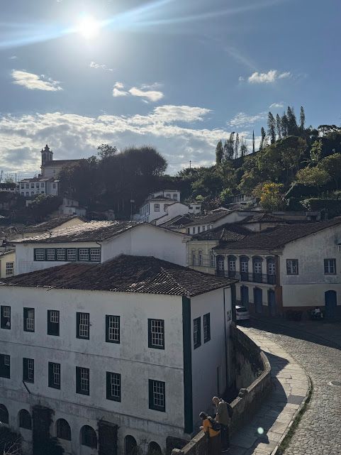 Edifícios brancos com telhados de telha alinham-se numa rua de paralelepípedos sob um céu ensolarado numa cidade na encosta de uma colina.