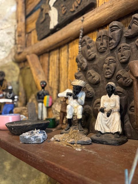 Expositor de prateleira com pequenas figuras esculpidas, incluindo pessoas sentadas e em pé, em frente a um fundo de madeira com entalhes de rostos.