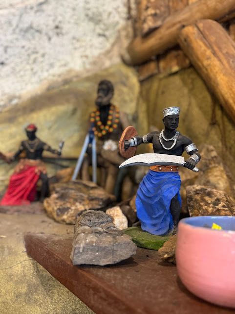 Miniaturas de figuras negras segurando armas e sentadas em um cenário com pedras e madeira.