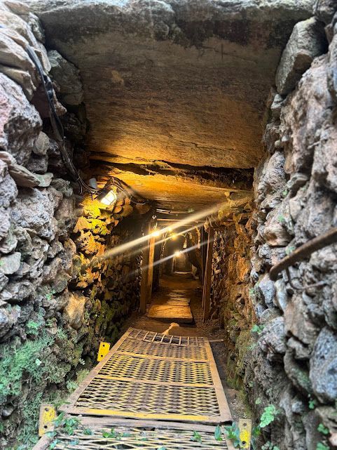 Túnel com paredes rochosas e passarela metálica, iluminado por luzes suspensas.