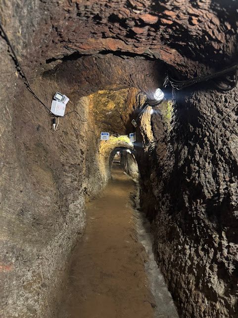 Túnel com paredes ásperas e marrons, iluminado por luzes suspensas, que se estende até o horizonte.