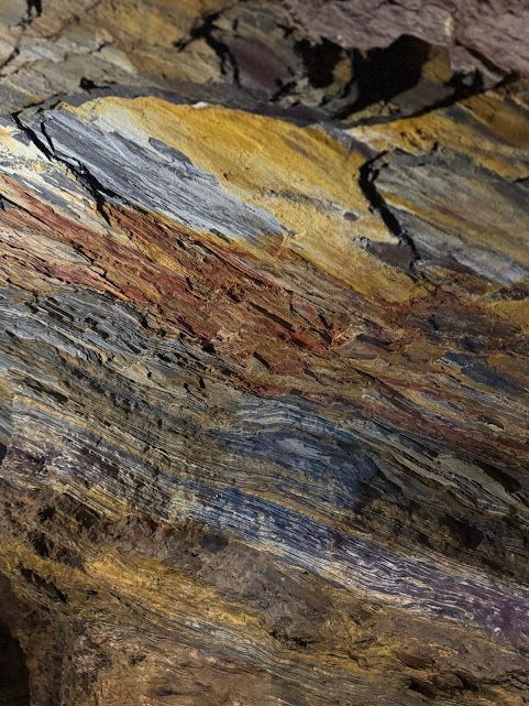 Detalhe de formações rochosas em camadas, em tons de dourado, cinza, marrom e vermelho.