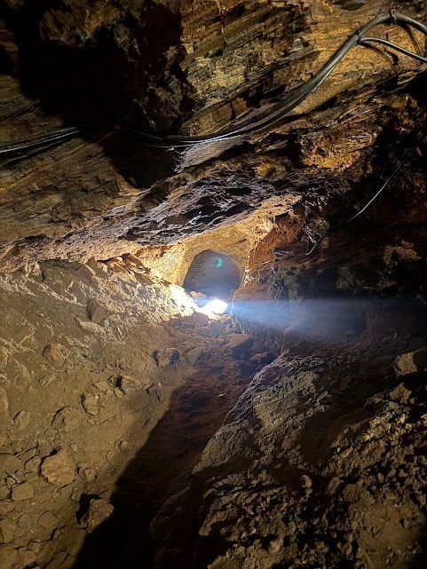 Interior da caverna com um feixe de luz brilhando através de uma abertura. Paredes e teto de rocha áspera e marrom.