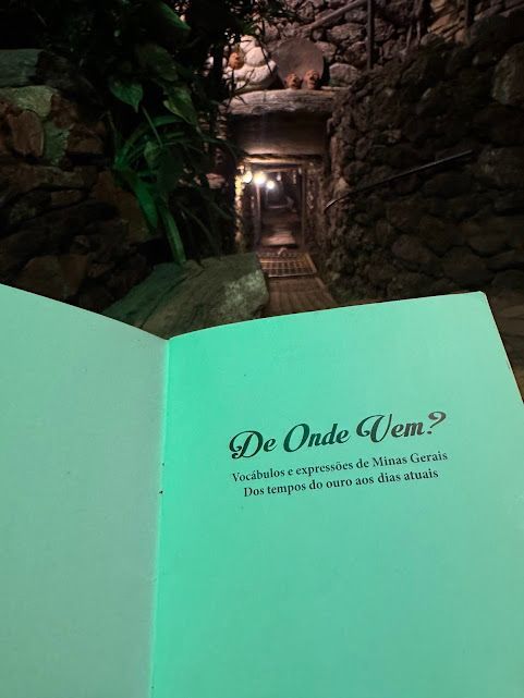 Livro aberto com texto em português, iluminado em luz verde, em frente a um túnel de pedra escuro.